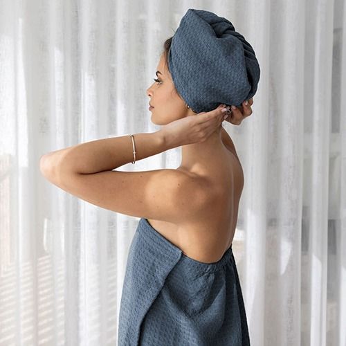 Woman in a textured towel wrap adjusts a matching towel on her head, standing in front of sheer white curtains
