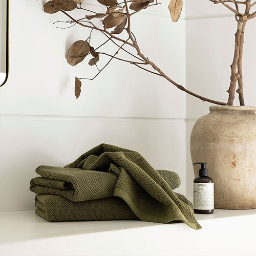 Olive green waffle-textured towels on a white surface beside a ceramic vase and a bottle of lotion