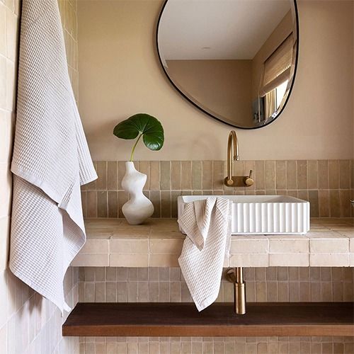 Modern bathroom sink area with a fluted white basin, gold faucet, abstract vase with green leaf, and textured beige towels