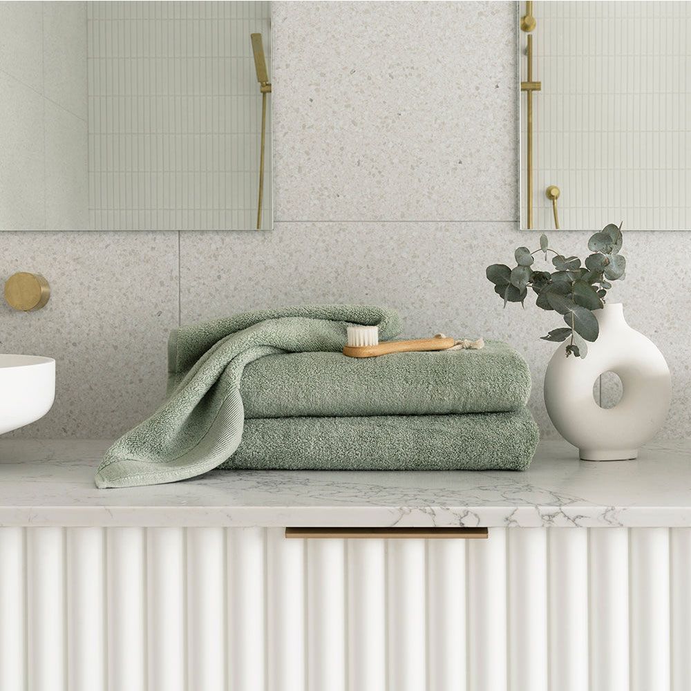 Soft green towels stacked on a marble bathroom counter with a toothbrush and vase of eucalyptus